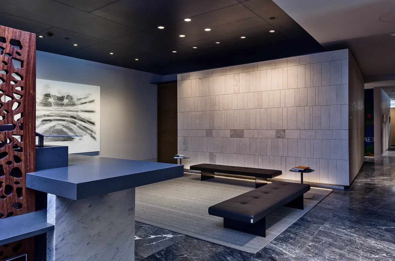 Recepción de oficinas con mostrador de piedra, muro de recubrimiento pétreo iluminado, bancas de piel y piso de mármol oscuro