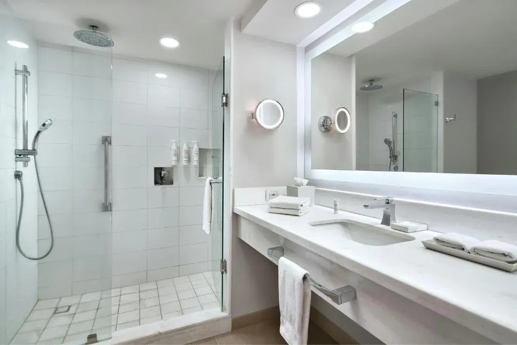 Baño de habitación de hotel con regadera de cristal, muros de azulejo blanco y cubierta de lavabo en piedra blanca con espejo iluminado