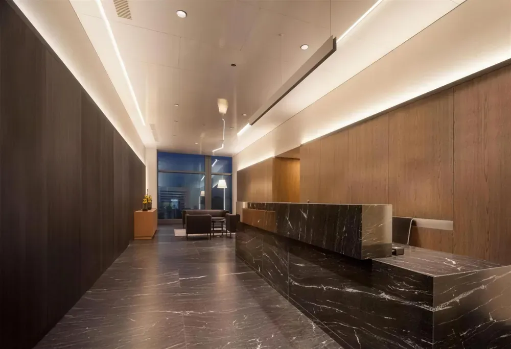 Recepción de oficinas con mostrador en mármol negro veteado, muros de madera e iluminación lineal
