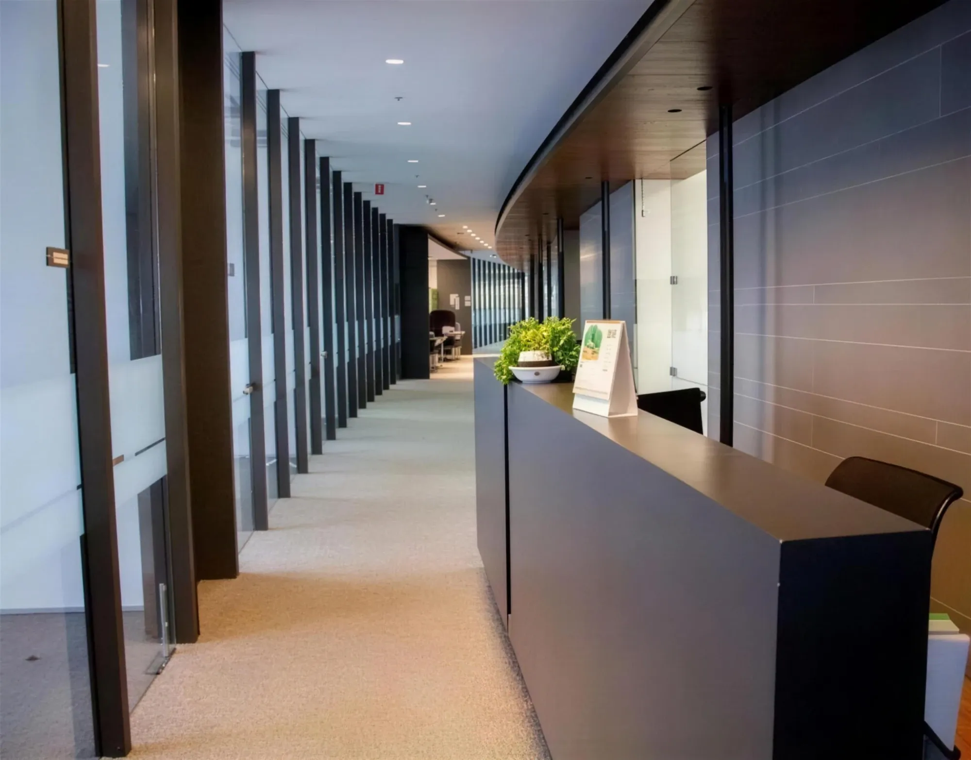 Pasillo de oficinas con muros de cristal, columnas oscuras y un mostrador de recepción integrado