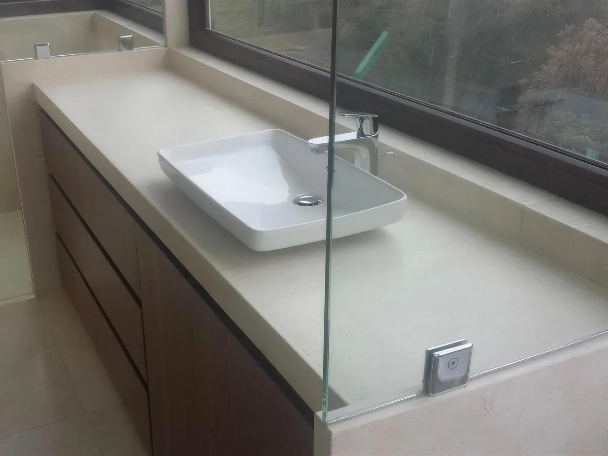 Mueble de baño en madera con cubierta de piedra beige, lavabo rectangular sobre cubierta y mampara de cristal